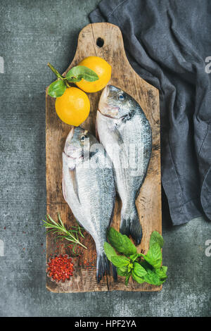 Frisches rohes Dorade oder Dorado Fisch mit Zitrone, Kräutern und Gewürzen auf rustikalem Holzbrett über graue konkreten Hintergrund, Ansicht von oben. Gesund, Diät, Stockfoto