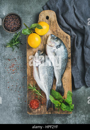 Frisches rohes Dorade oder Dorado Fisch mit Zitrone, Kräuter und Gewürze in Schalen auf rustikalem Holzbrett über graue konkreten Hintergrund, Ansicht von oben. Gesund, Stockfoto