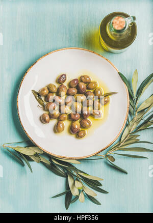 Eingelegte grüne mediterrane Oliven auf weißen Keramikplatte, Olivenbaum Zweig und natives Olivenöl in Glasflasche über Licht blau bemalte hölzerne backg Stockfoto
