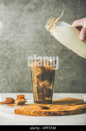 Hand des Mannes bereit, Milch aus der Flasche, Glas mit Eiswürfeln gefroren Kaffee auf Olivenholz und Marmor dienen Board über graue Tisch, dunkle Sperrholz w gießen Stockfoto