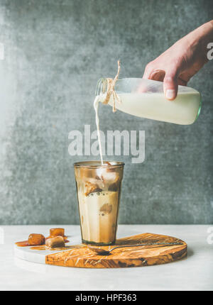 Mannes Hand Gießen Milch Iced Caramel Latte Kaffee mit Eiswürfeln gefroren Kaffee im Glas servieren Holz und Marmor Board über graue Tisch cocktail, Stockfoto