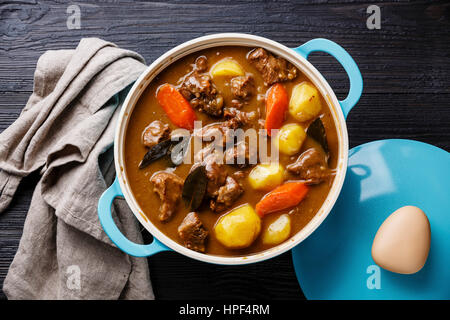 Rindfleisch geschmort mit Kartoffeln, Möhren und Gewürzen in gusseisernen Pfanne auf verbrannte schwarze Holz Hintergrund Stockfoto