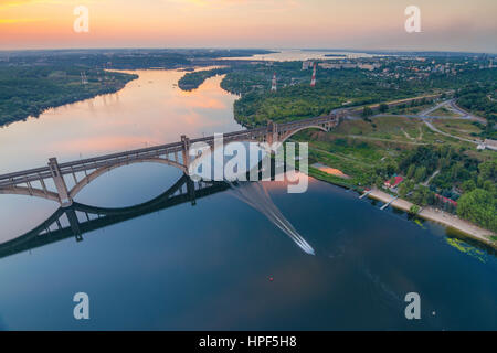 Abends auf Dnjepr und Preobraschenskij gewölbte Brücke aus einer Höhe von 180m mit Motorboot im Wasser, Zaporozhye, Ukraine Stockfoto