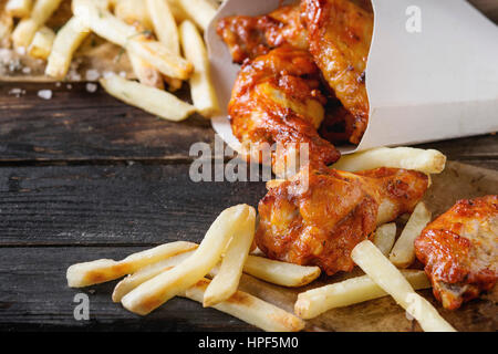 Fast-Food gebraten Huhn Beine, Flügel und Pommes frites Kartoffeln in Lunch-Boxen mit Salz und Ketchup serviert auf Backpapier über alte dunkle wo Soße Stockfoto