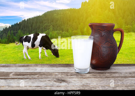 Milch im Glas und Ton Krug auf alten Holztisch mit grasende Kuh auf der Wiese mit Sonne im Hintergrund. Topf und Glas Milch auf Holztisch Stockfoto