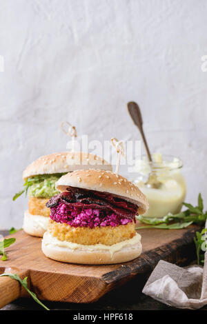 Zwei vegetarische Hamburger mit Zwiebeln und Käse Schnitzel, Avocado-Salat, Rucola, gebratene rote Bete und Joghurt Sauce auf hölzernen Portion Board über Textur Stockfoto