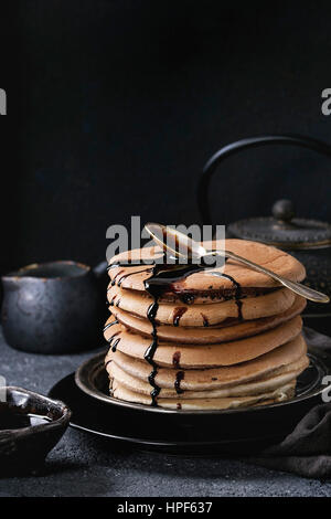 Stapel von hausgemachten amerikanischen Ombre Schokolade Pfannkuchen mit Johannisbrot-Honigsoße serviert auf schwarze Platte mit Krug mit Sahne und Teekanne über schwarze Stein Textur Stockfoto