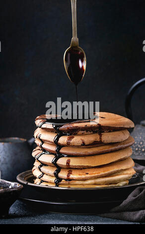 Stapel von hausgemachten amerikanischen Ombre Schokolade Pfannkuchen mit Johannisbrot-Honig-Sauce, vom Löffel fließt auf schwarze Platte mit Sahne und Teekanne Ove Krug serviert Stockfoto