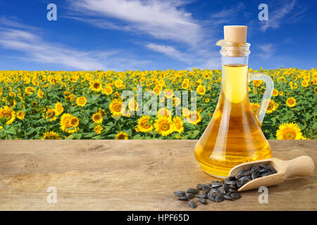 Sonnenblumenöl in Glasflasche und Samen auf Holztisch mit Sonnenblumenfeld im Hintergrund. Sonnenblumenfeld bei blauem Himmel. Foto mit Textfreiraum Stockfoto