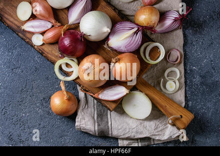 Vielzahl von ganze und geschnittene Schalotte, gelb, rot und weiß Zwiebeln auf Schneidbrett aus Holz schneiden auf Textile Serviette über dunkle Stein Textur Zeitmessung Stockfoto