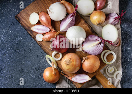 Vielzahl von ganze und geschnittene Schalotte, gelb, rot und weiß Zwiebeln auf Schneidbrett aus Holz schneiden auf Textile Serviette über dunkle Stein Textur Zeitmessung Stockfoto