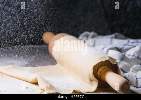 Frische hausgemachte Teig für Nudeln Tagliatelle auf Holz Nudelholz mit Sieb Prise Mehl und Küche Handtuch über dunklen grauen Tabellenhintergrund gerollt. Stockfoto
