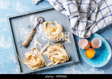 Frisches rohes hausgemachte verdrehten Teigwaren Tagliatelle mit Eigelb und Pasta Eierschneider Holztablett mit Küchentuch über leichte blaue konkrete backgr Stockfoto