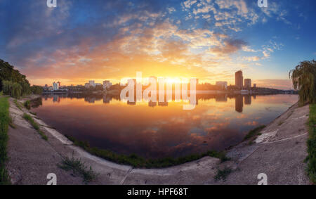 Schöne Stadtbild bei Sonnenuntergang. Erstaunliche Panoramasicht auf die Stadt vom Ufer Flusses. Bunte blauer Himmel mit Wolken und Sonne spiegelt sich im Wasser Stockfoto