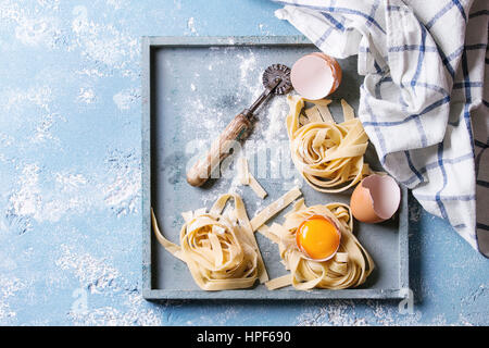 Frisches rohes hausgemachte verdrehten Teigwaren Tagliatelle mit Eigelb, Schale und Pasta Eierschneider Holztablett mit Küchentuch über blaue Leichtbeton Stockfoto