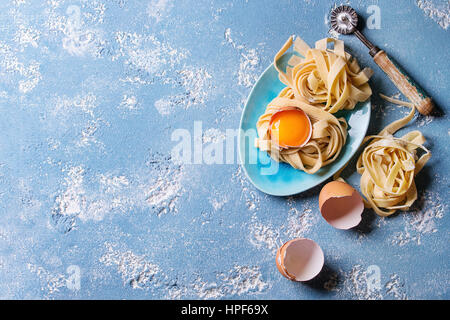 Frisches rohes hausgemachte verdrehten Teigwaren Tagliatelle mit Eigelb, Schale und Pasta Eierschneider Holztablett mit Küchentuch über blaue Leichtbeton Stockfoto