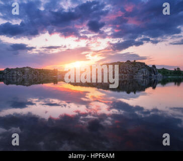 Fluss gegen bunten Himmel mit Wolken und Felsen bei Sonnenuntergang im Sommer. Wunderschöne Landschaft mit See, Berge, Sonne und blauen Wolkenhimmel Stockfoto