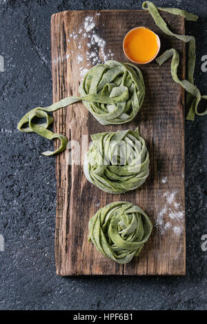 Frischen rohen ungekochten hausgemachte verdrehte Grüne Spinat Pasta Tagliatelle mit Eigelb und Mehl auf Schneidbrett aus Holz schneiden über dunkle Textur concret Stockfoto
