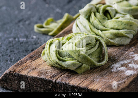 Frischen rohen ungekochten hausgemachte verdrehte Grüne Spinat Pasta Tagliatelle mit Mehl auf Schneidbrett aus Holz schneiden über konkrete Hintergrund dunkle Textur. Stockfoto