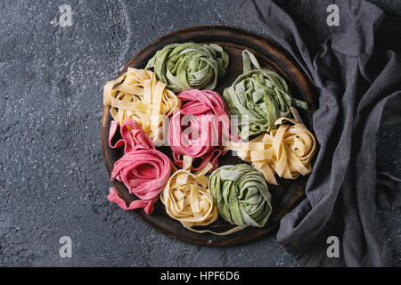 Verschiedene farbige frische rohe hausgemachte Teigwaren Tagliatelle grün Spinat, rosa rote Beete und gelb auf Holzbrett mit Textil über dar Stockfoto