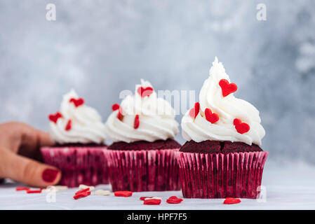 Red Velvet Cupcakes mit Buttercreme frosting und verziert mit Herzen geformt Streuseln.  Eine Frau ist einer von ihnen greifen. Stockfoto