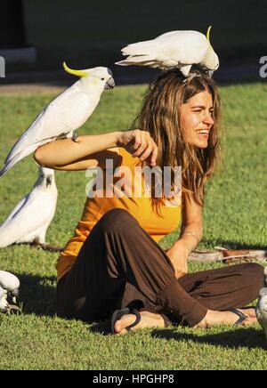 Hübsche junge Frau mit Kakadu Vögel balancieren auf ihren Schultern sitzen auf dem Rasen des Botanischen Gartens in Sydney Australia Stockfoto