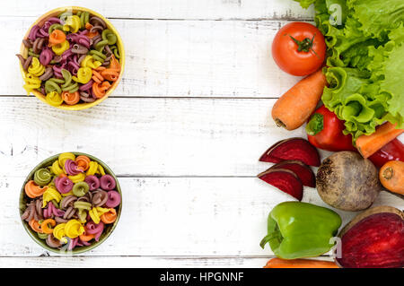 Bunte Nudeln in eine Schüsseln auf weißem Hintergrund aus Holz, mit frischem Gemüse (Rüben, Grüns, Karotten, Tomaten, Paprika). Gesunde Ernährung-Konzept. Stockfoto