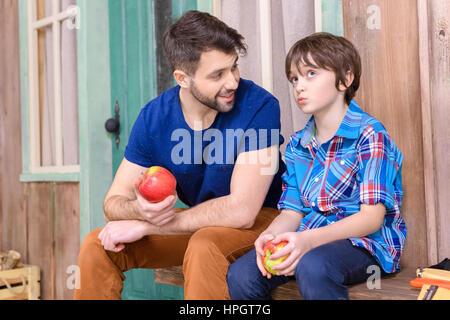 Vater und niedlichen nachdenklich Sohn sitzen auf Holzbank und Verzehr von Äpfeln Stockfoto