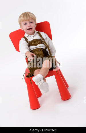 Kleiner Junge in Lederhose Sitzt Auf Rotem Stuhl - kleiner Junge in Lederhose sitzt auf einem roten Stuhl, Modell veröffentlicht Stockfoto