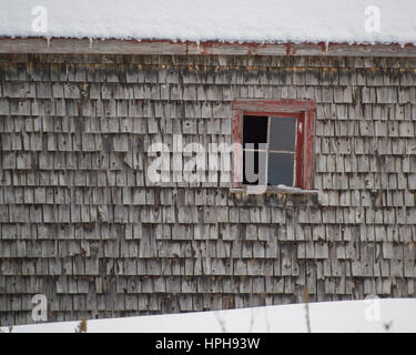 Alten verwitterten Zeder schüttelt Schindeln Scheune mit roten Fenster Winter. Schnee auf dem Dach und Boden. Stockfoto