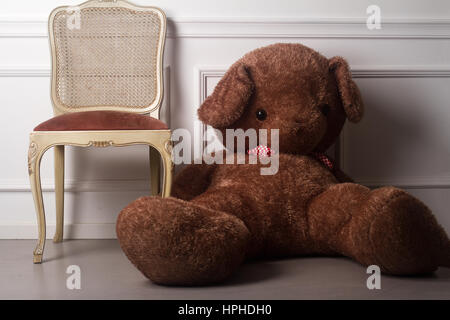 horizontale Vorderansicht eines großen braunen Teddybären sitzen auf dem Boden neben einem Luxus Stuhl gegen eine weiße Wand Stockfoto