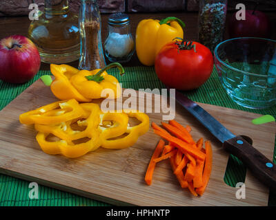 Große gelbe saftige Scheiben Paprika Ringe auf einem Holzbrett auf einem Küchentisch mit Karotten, rote Tomaten, Äpfel, Pflanzenöl, Salz, Gewürze Stockfoto