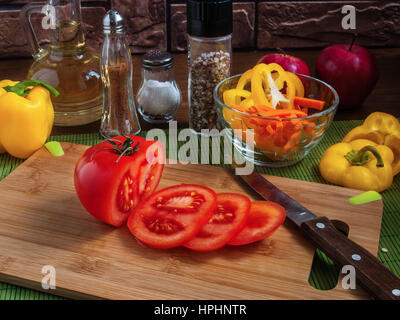 Große gelbe saftige Scheiben Paprika Ringe auf einem Holzbrett auf einem Küchentisch mit Karotten, rote Tomaten, Äpfel, Pflanzenöl, Salz, Gewürze Stockfoto