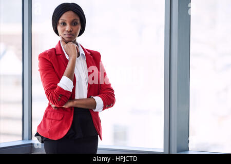 Zuversichtlich Geschäftsfrau mit Fett Körpersprache während des Tragens eines roten Blazer mit großen Fenstern hinter ihr im Hintergrund in die Kamera schaut. Stockfoto