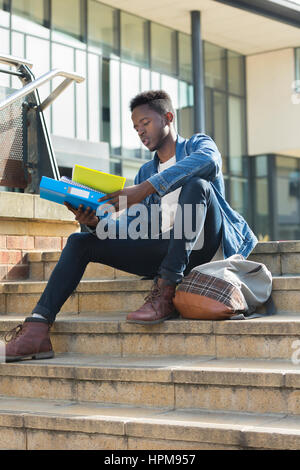 Ein junger Mann sitzt auf einige Schritte in der Stadt, wie er studiert. Er hält seine Bücher und seine Tasche ist an den Füßen. Stockfoto