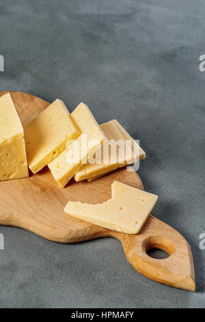 Harter Käse in Scheiben geschnitten auf einem Holzbrett zu Hause. Käse zum Mittagessen. Grauer Hintergrund im Hintergrund. Der Käse hat kleine Löcher. Eines der Stücke von Käse Stockfoto