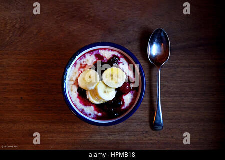 eine Schüssel Brei mit rote Himbeer Frucht und Banane in ein weißes Metall Schüssel mit einem blauen Rand einer einzigen Silberlöffel daneben auf einem Holztisch, t Stockfoto