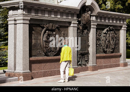Frau mit gelber Jacke und Tasche schaut das Denkmal für Alexander 1 Alexandrovsky Garden in Moskau. Park mit Rasenflächen, Sommer Blüten & mehrere memo Stockfoto