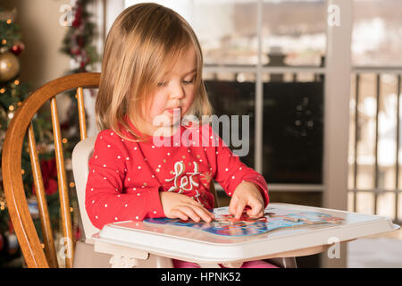 Junge weibliche Babymädchen in einen Hochstuhl, ein Puzzle auf einer Plastikschale zu Weihnachten mit Baum im Hintergrund Stockfoto