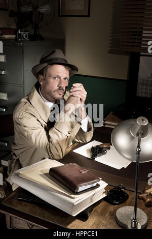 Zuversichtlich Detektiv Rauchen am Schreibtisch im Trenchcoat, der 1950er Jahre Film-Noir-Stil. Stockfoto