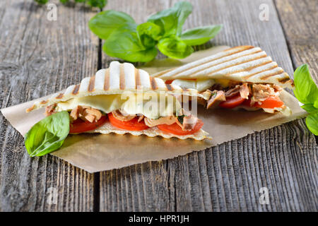 Getoastete Panini mit Thunfisch, Tomaten, geschmolzenem Mozzarella und frischem Basilikum auf Butterbrotpapier auf einem Holztisch serviert Stockfoto