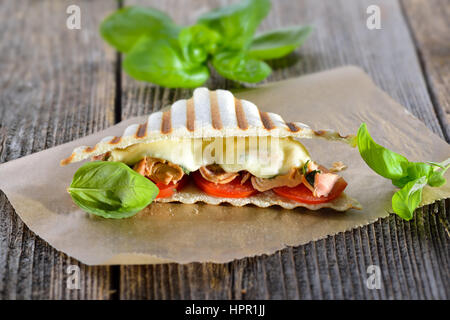 Getoastete Panini mit Thunfisch, Tomaten, geschmolzenem Mozzarella und frischem Basilikum auf Butterbrotpapier auf einem Holztisch serviert Stockfoto