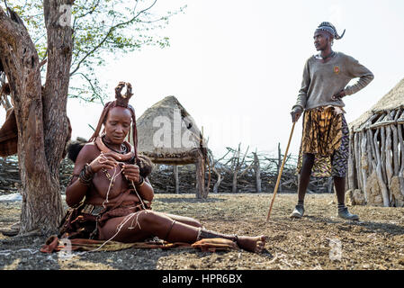 Himba Frau weben Kunststoff-Fäden aus alten Plastiktüten. Junger Himba Mann mit traditionellen Scorpion Endstück im Hintergrund. Stockfoto