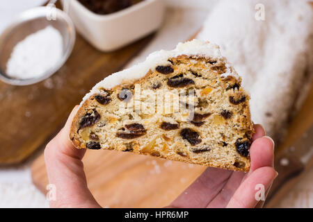 Zarte Frauenhand hält ein Stück Weihnachten Christstollen, Brot und Zutaten im Hintergrund, Ansicht von oben, Nahaufnahme Stockfoto