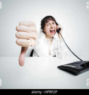 eine feindliche Frau zeigt Daumen nach unten gegen uns (an der Kamera) beim Telefonieren an ihrem Schreibtisch Stockfoto