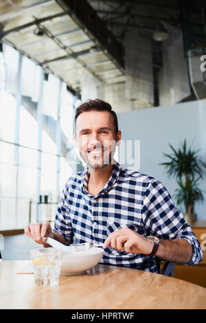 Gut aussehender Mann im Café Essen mit Gabel und Messer in Händen lächelnd und Blick in die Kamera Stockfoto