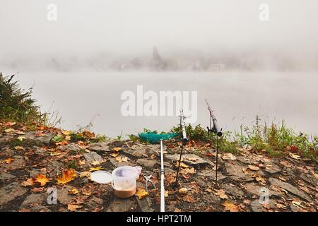 Angelruten, Kescher und Köder Angeln am Ufer Flusses. Dichter Nebel während Herbstmorgen in der schönen Natur in der Tschechischen Republik Stockfoto