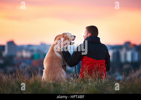 Sonne genießen. Mann ist gelbe Labrador Retriever streicheln. Junger Mann sitzt auf dem Hügel mit seinem Hund. Wunderbaren Sonnenaufgang in der Stadt. Prag in Tschechien Stockfoto