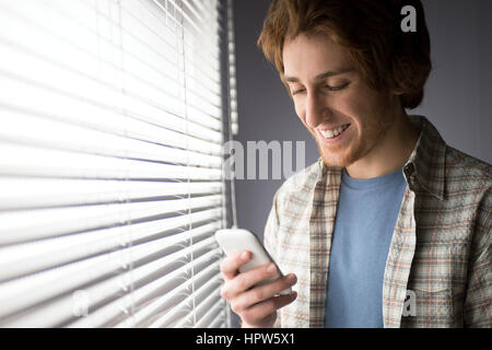 Junger Mann lächelnd und mit einem Smartphone vor einem Fenster. Stockfoto