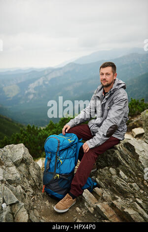 Porträt eines Mannes mit Wanderausrüstung sitzt auf einem Felsen bewundern Sie die Aussicht von oben auf einem Berg während Sie Wandern in der Wildnis Stockfoto